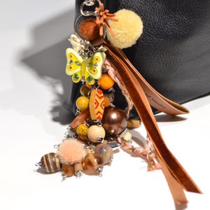 Puede incluir: Un colgante decorativo para bolso con una figura de mariposa amarilla con detalles verdes, varias cuentas marrones y amarillas, un pompón amarillo difuso y largas tiras de cuero marrón, unido a un mosquetón de metal plateado en un bolso negro.