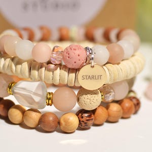 Può includere: Una pila di quattro braccialetti di perline con perline rosa cipria opaco, crema e legno naturale. Il set include un ciondolo rotondo in metallo dorato inciso con la parola STARLIT, perline in pietra lavica porosa e una perlina di cristallo sfaccettato.