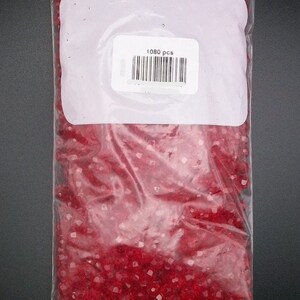 May include: A clear plastic bag filled with 1080 red, faceted glass beads. The bag has a white label with the text "1080 pcs" and a barcode.