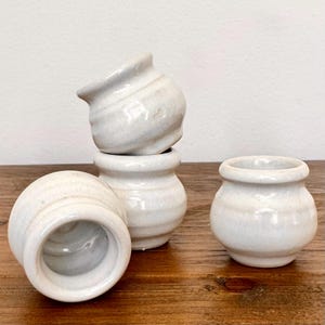 May include: Four small, white ceramic pots with a textured, marbled appearance. The pots are stacked and arranged on a wooden surface, showcasing their rounded bodies and open tops. The neutral color and simple design suggest a minimalist aesthetic.