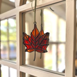 May include: A stained glass maple leaf ornament featuring vibrant red and orange glass segments held together by dark metal solder lines. The leaf is suspended by a thin green ribbon, showcasing a detailed vein pattern throughout the glass.