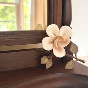 May include: A dark brown wooden mirror frame featuring a decorative brass branch accent with textured leaves and a single cream-colored ceramic rose flower attached to the lower right corner of the frame, reflecting a window in the glass.