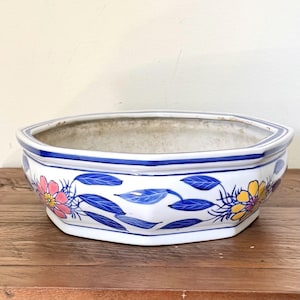 May include: White ceramic planter with a blue rim and floral design. The planter has a hexagonal shape and is decorated with pink and yellow flowers and blue leaves. The planter sits on a wooden surface.