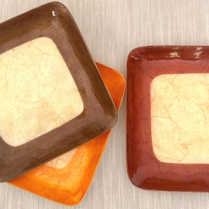 May include: A close-up shot of three square decorative trays. The trays have a brown, orange, and reddish-brown border with a cream-colored center. The trays are stacked and arranged on a neutral-colored surface.