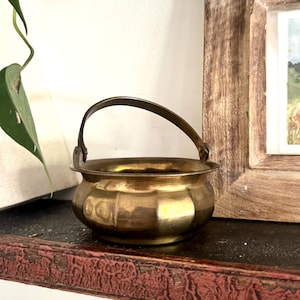 May include: A small, antique brass pot with a curved handle. The pot has a rounded body with vertical indentations and sits on a dark surface. A framed picture and a green plant are in the background.