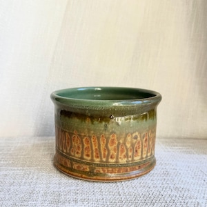 May include: A cylindrical ceramic bowl featuring a smooth, glossy sage green glaze on the interior and upper rim, transitioning into a textured, mottled brown and tan drip pattern across the lower exterior body of the vessel.