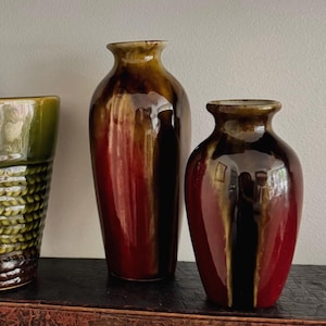 May include: Two ceramic vases with a glossy finish featuring a deep red base color that transitions into a dark brown glaze at the neck and rim. The vases have a rounded body and a narrow opening, displaying a smooth, reflective surface texture.