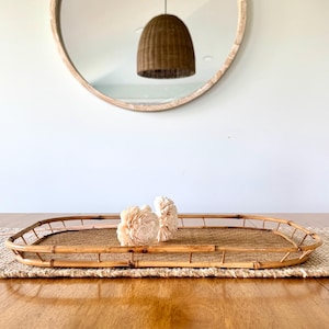 May include: A rectangular bamboo tray with a woven base and a raised border. Two cream-colored decorative flowers rest on the tray. The tray sits on a woven placemat on a wooden table. A round mirror and a woven pendant light are in the background.