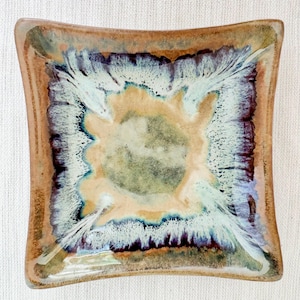 May include: A square ceramic dish featuring a reactive glaze with a central tan and olive green core, surrounded by a vibrant white and blue feathered pattern, framed by a dark brown and tan border with slightly curved edges.