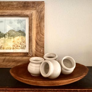 May include: A wooden framed watercolor painting of a landscape scene. Four small, off-white ceramic pots are arranged on a dark wooden oval-shaped bowl. The pots have a rounded shape with a small opening at the top.