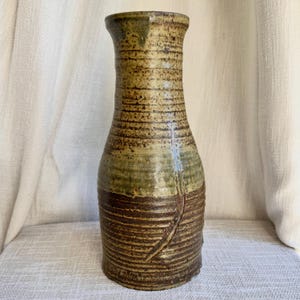 May include: A tall, cylindrical ceramic vase featuring a textured, ribbed surface. The upper portion is finished in a mottled olive-green glaze, while the lower section displays a deep, earthy brown tone with a prominent vertical incised line.