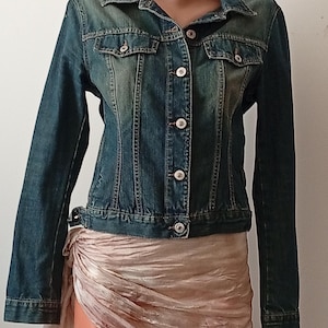 May include: A blue denim jacket with a classic collar, button-up front, and two chest pockets. The jacket has a slightly faded appearance and is paired with a draped, light-coloured skirt. The jacket features white buttons and a button closure at the waist.
