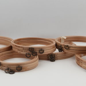 Puede incluir: Una colección de aros de bordado de madera de varios tamaños. Los aros son de color marrón claro con herrajes metálicos y un detalle tejido marrón oscuro. Algunos aros tienen un pequeño cierre metálico. Los aros están apilados.