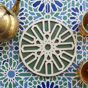 Puede incluir: Una tetera de latón y dos vasos de té con hojas de menta están dispuestos sobre una superficie de baldosas estampadas. En el centro, un posavasos decorativo redondo, blanco y verde, con un intrincado diseño geométrico. El fondo es un patrón de baldosas con estrellas azules y verdes.