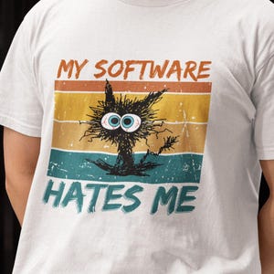 Puede incluir: Camiseta blanca con un gráfico desgastado. El diseño presenta el texto "MY SOFTWARE HATES ME" y un gato de dibujos animados con ojos grandes. El gráfico utiliza una combinación de colores retro de naranja, amarillo, verde azulado y azul.
