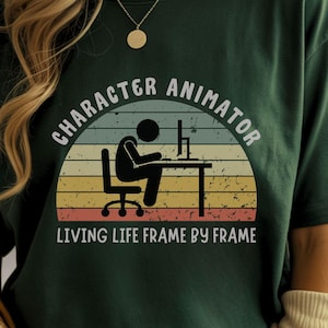 Op de afbeelding: Donkergroen t-shirt met een retro ontwerp met de tekst "Character Animator" en een stokfiguur die op een computer werkt. De tekst "Living Life Frame by Frame" staat eronder.