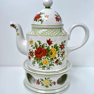 Op de afbeelding: Witte keramische theepot met een bloemmotief in rood, geel en oranje. De theepot heeft een bijpassende deksel en staat op een bijpassende warmer. Het ontwerp bevat groene accenten en een geometrisch patroon.
