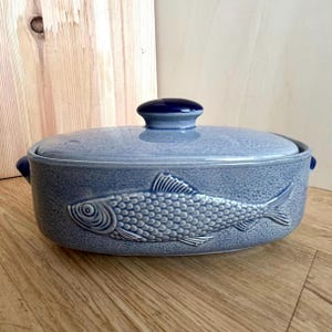 Könnte beinhalten: Hellblaue Keramik-Auflaufform mit Deckel. Die Schüssel hat eine strukturierte Oberfläche und ein erhabenes Relief eines Fisches an der Seite. Der Deckel hat einen dunkelblauen Knopf. Die Schüssel steht auf einer Holzoberfläche.