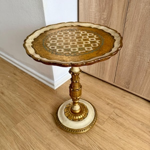 Puede incluir: Una pequeña y ornamentada mesa auxiliar con una tapa redonda y una base decorativa. La mesa presenta una combinación de colores dorados y crema con intrincados detalles en la tapa y la base. La tapa tiene un borde dorado y un diseño.