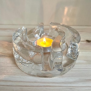 May include: A clear glass candle holder with a textured surface. The holder has a circular design with curved sections. A lit tealight candle sits in the center, casting a warm glow. The candle holder is on a wooden surface.