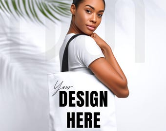 May include: White tote bag with black straps featuring the text "Your DESIGN HERE" in a bold, black font. The bag is held by a person wearing a white t-shirt. The background is a soft white with a palm leaf shadow.
