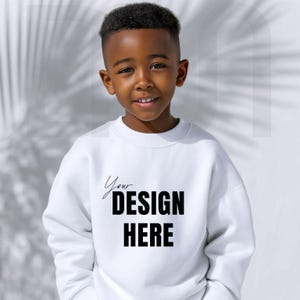 May include: A young boy wearing a white sweatshirt with the text "Your DESIGN HERE" in black. The sweatshirt is a crew neck style. The background is a light gray with shadows.