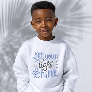May include: A young person wearing a white sweatshirt with the text "Let your light Shine" in blue and black script. The sweatshirt has a crew neck and long sleeves. The background is a soft, neutral color.