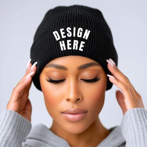 May include: Black beanie with the text "DESIGN HERE" in white block letters. The beanie is ribbed and appears to be made of a soft material. The beanie is worn by a person with their eyes closed and hands on their head.