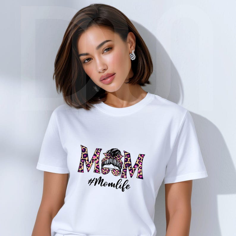 May include: White t-shirt featuring the word "MOM" in a leopard print design with a bun graphic. Below the word "MOM" is the hashtag "#Momlife" in a script font. The shirt is short-sleeved.