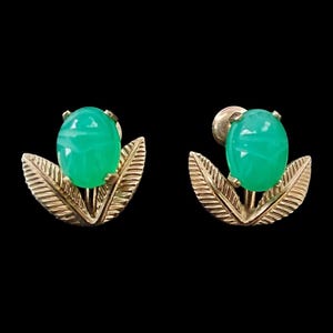 Puede incluir: Un par de pendientes vintage con piedras preciosas ovaladas de color verde brillante engastadas en metal dorado. Cada pendiente tiene un diseño en forma de hoja debajo de la piedra preciosa, creando un tema floral o botánico. El fondo es negro.