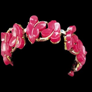 May include: A vintage bracelet featuring a floral design. The bracelet has gold-toned metal links and is adorned with vibrant, fuchsia-colored flower-shaped elements. The flowers are intricately carved, adding depth and dimension to the piece. The bracelet is set against a black background.