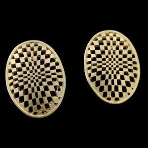 May include: Two gold-colored, oval-shaped earrings. Each earring features a checkered pattern of open squares, creating a lattice effect. The earrings are set against a black background, highlighting their intricate design.
