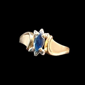 May include: A gold ring featuring a central marquise-cut blue gemstone flanked by small clear stones. The band has a smooth, polished finish that tapers toward the sides, creating a bypass-style setting that highlights the deep blue center stone.