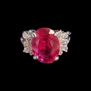 Op de afbeelding: Een zilveren ring met een grote, ovaal geslepen robijn. De robijn heeft een heldere rode kleur en is omgeven door kleinere, heldere edelstenen. De ring staat tegen een zwarte achtergrond.