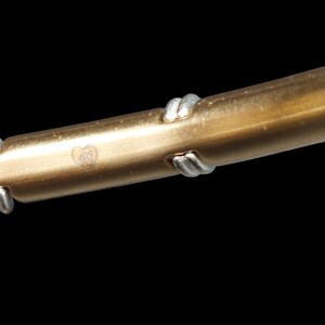 K&ouml;nnte beinhalten: Nahaufnahme eines goldfarbenen Metallarmbands mit silberfarbenen Akzenten. Das Armband hat ein herzf&ouml;rmiges Emblem mit den Initialen "GB". Das Armband ist vor einem schwarzen Hintergrund platziert.