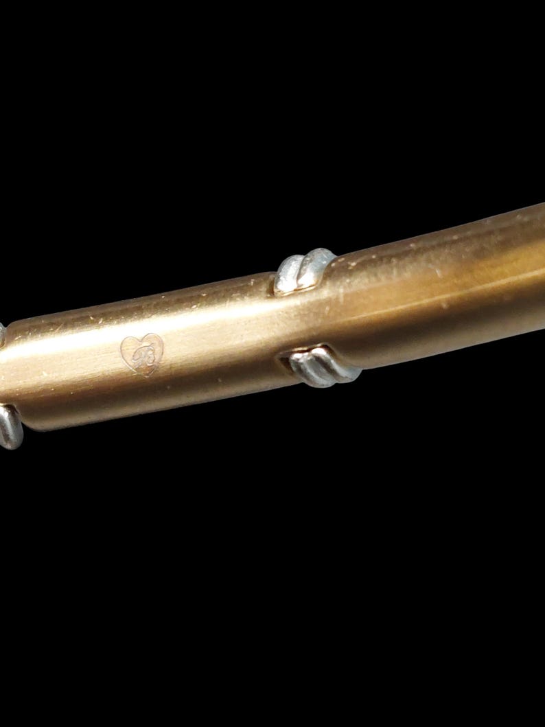 K&ouml;nnte beinhalten: Nahaufnahme eines goldfarbenen Metallarmbands mit silberfarbenen Akzenten. Das Armband hat ein herzf&ouml;rmiges Emblem mit den Initialen "GB". Das Armband ist vor einem schwarzen Hintergrund platziert.