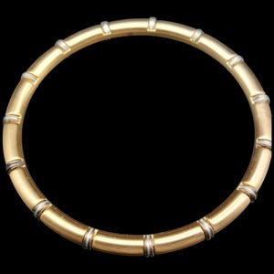 K&ouml;nnte beinhalten: Eine goldfarbene Halskette mit Bambus-Design. Die Halskette weist abwechselnde Abschnitte aus gold- und silberfarbenem Metall auf, wodurch ein segmentiertes Aussehen entsteht. Der Schmuck ist vor einem schwarzen Hintergrund platziert.