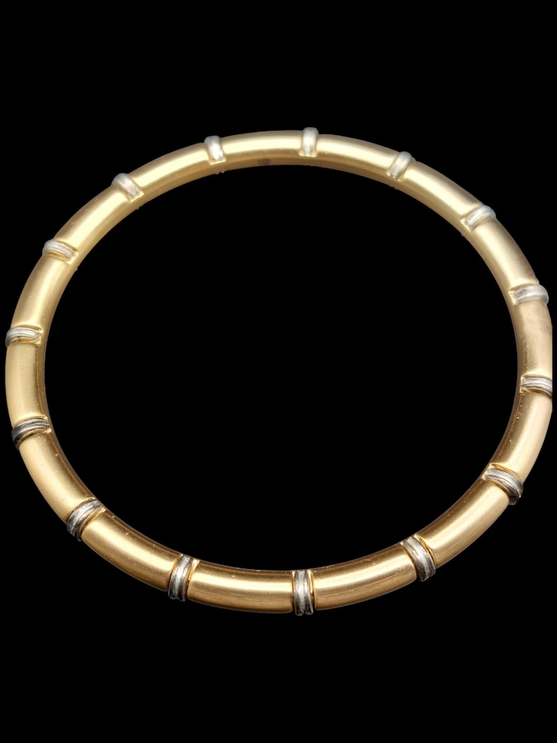 K&ouml;nnte beinhalten: Eine goldfarbene Halskette mit Bambus-Design. Die Halskette weist abwechselnde Abschnitte aus gold- und silberfarbenem Metall auf, wodurch ein segmentiertes Aussehen entsteht. Der Schmuck ist vor einem schwarzen Hintergrund platziert.