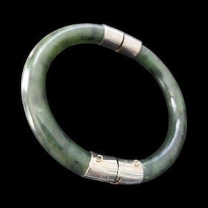 Könnte beinhalten: Ein grünes Jade-Armreif-Armband mit einem silbernen Verschluss. Das Armband hat eine tiefgrüne Farbe mit Farbvariationen. Der silberne Verschluss ist rechteckig und verbindet die beiden Enden. Der Hintergrund ist schwarz.