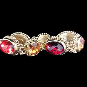 May include: A gold-toned bracelet featuring a series of colorful gemstones. The bracelet showcases a variety of shapes, including oval and rectangular stones in shades of red, orange, and yellow. The metalwork has an ornate, textured design.