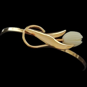 May include: A gold-toned metal bangle bracelet featuring a stylized tulip design. The bracelet has a smooth, curved band that loops around a central gold leaf element, which is attached to a carved, off-white tulip-shaped floral ornament.