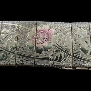 May include: Close-up of a silver-toned bracelet with a floral design. The bracelet features a repeating pattern of flowers with pink centers and detailed leaves. The background is black.