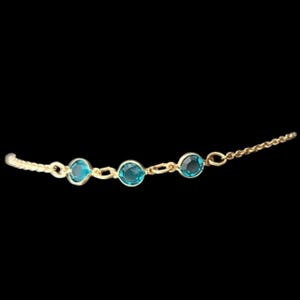 May include: A gold-toned chain bracelet featuring three round, bezel-set teal blue stones spaced evenly along the center. The delicate metal chain links connect each stone setting, creating a continuous, refined piece of jewelry against a black background.