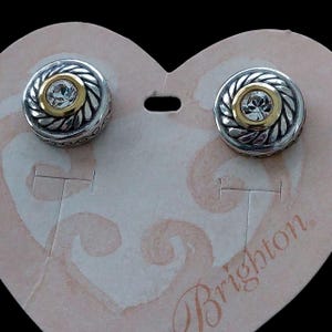 May include: A pair of silver-toned stud earrings with a gold-colored center and a clear, round gemstone. The earrings have a decorative, rope-like border and are displayed on a heart-shaped card.
