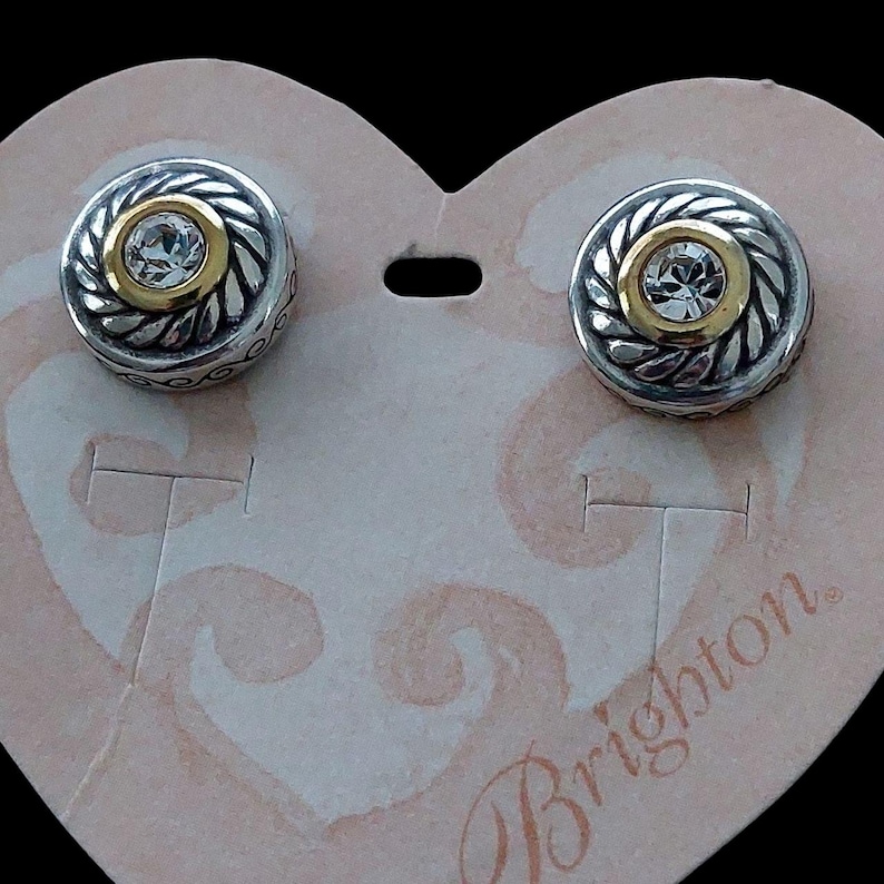 May include: A pair of silver-toned stud earrings with a gold-colored center and a clear, round gemstone. The earrings have a decorative, rope-like border and are displayed on a heart-shaped card.