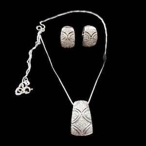 May include: A silver jewelry set featuring a pendant necklace and matching hoop earrings. The pendant and earrings are adorned with a repeating geometric pattern and small, sparkling embellishments. The jewelry is displayed against a black background.