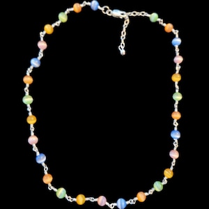 Puede incluir: Un collar de cuentas multicolores con una cadena y cierre plateados. El collar presenta cuentas redondas en azul, naranja, amarillo, rosa y verde. Las cuentas están ensartadas con alambre plateado.