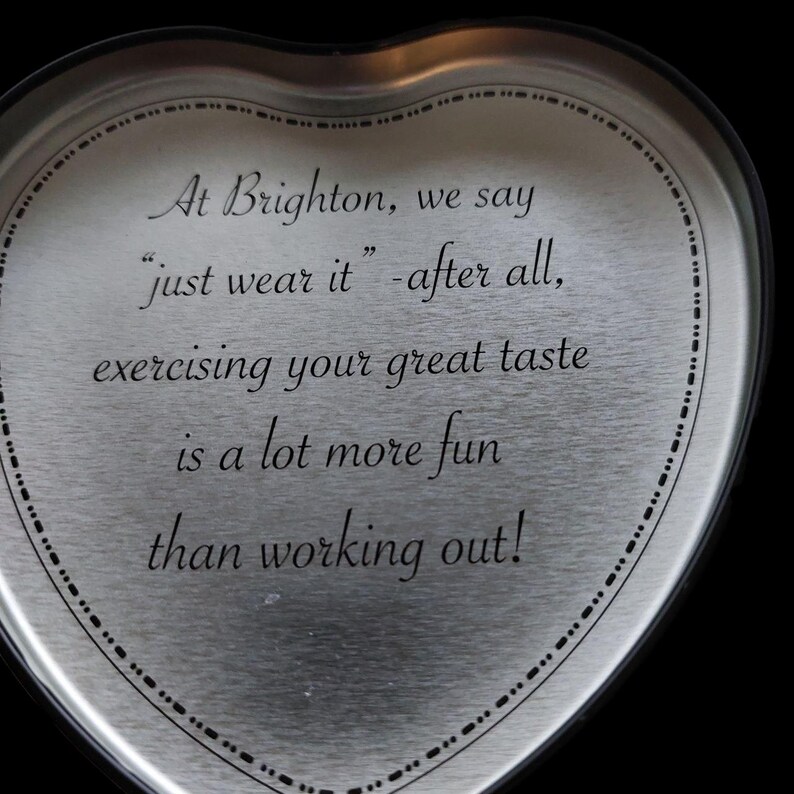 May include: A heart-shaped, silver-colored metal container with a black border. The container has the text "At Brighton, we say 'just wear it' - after all, exercising your great taste is a lot more fun than working out!"
