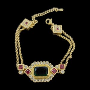 May include: A gold-tone bracelet featuring a central rectangular dark green gemstone surrounded by small clear stones. The design includes square red gemstones and additional clear stones along the double chain, finished with a lobster claw clasp.