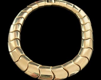 May include: A gold-toned necklace with a snake-like design. The necklace is composed of interlocking, curved segments that create a flexible, collar-style piece. The jewelry is set against a black background.